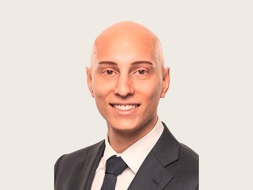 Anastasios Kapetanos, Claims Underwriter bei Hiscox, trägt einen dunklen Anzug, ein weißes Hemd und eine dunkle Krawatte vor hellem Hintergrund.