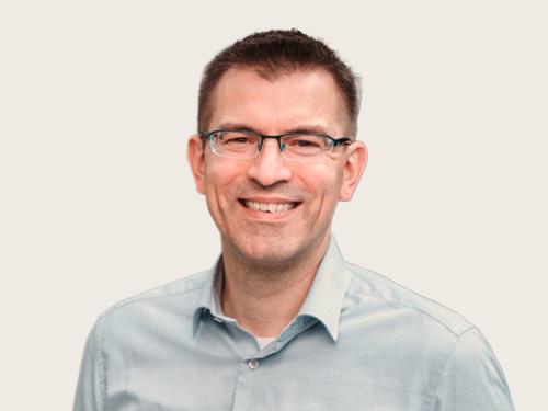 Ein lachender Mann mit Brille und einem hellblauen Hemd, dessen oberer Knop geöffnet ist, vor einem hellen Hintergrundblauen Hemd vor einem neutralen Hintergrund.