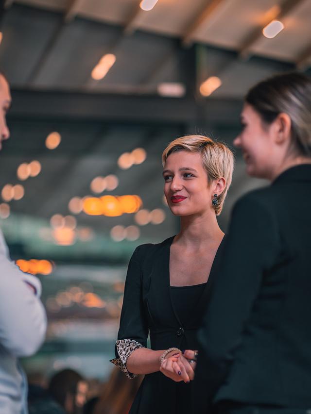 Eine hübsche blonde Frau mit kurzen Haaren lächelt zwei Gesprächspartnern auf einer Messe zu