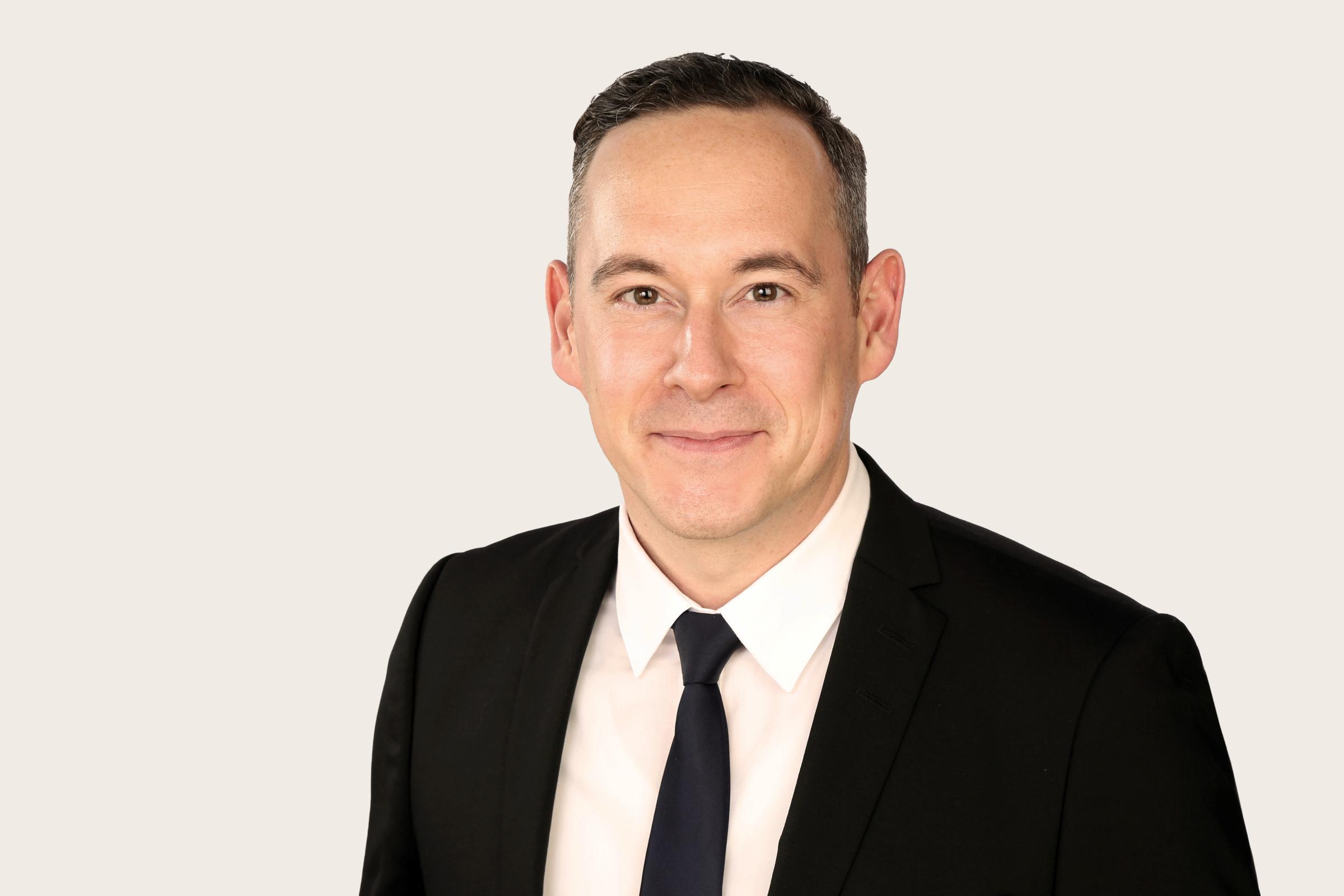 Patrick Klawitter hat kurzes dunkles Haar, trägt einen schwarzen Anzug, eine schwarze Krawatte und ein weißes Hemd. Er lächelt in die Kamera.