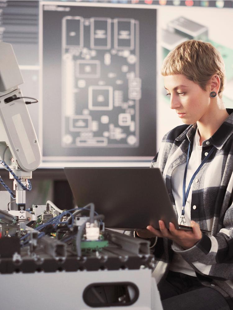 Eine junge blonde Frau mit kurzen Haaren sitzt vor einer Werkbank mit elektronischen Geräten und einem Roboterarm. Sie hält einen Laptop in der Hand über ihrem Schoß, in den sie hineinschaut. Im Hintergrund sieht man verschwommen einen großen Bildschirm mit technischen Zeichnungen.