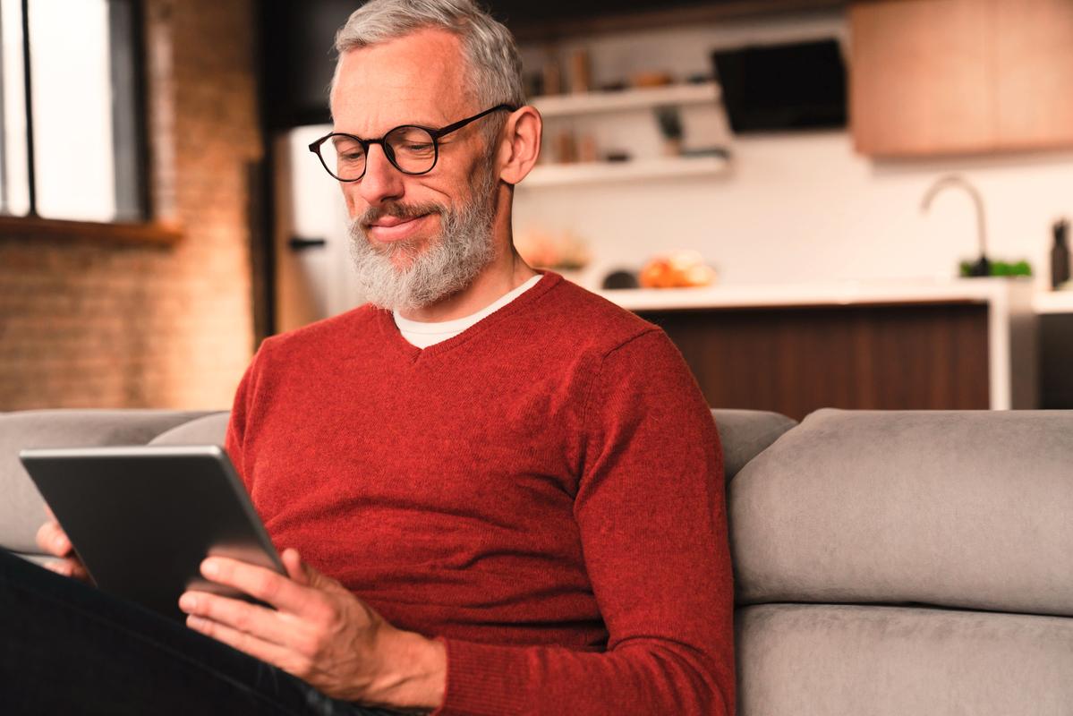 Eine ältere Person mit grauen Haaren und Bart, trägt eine Brille und einen roten Pullover, sitzt auf einem grauen Sofa und hält ein Tablet.