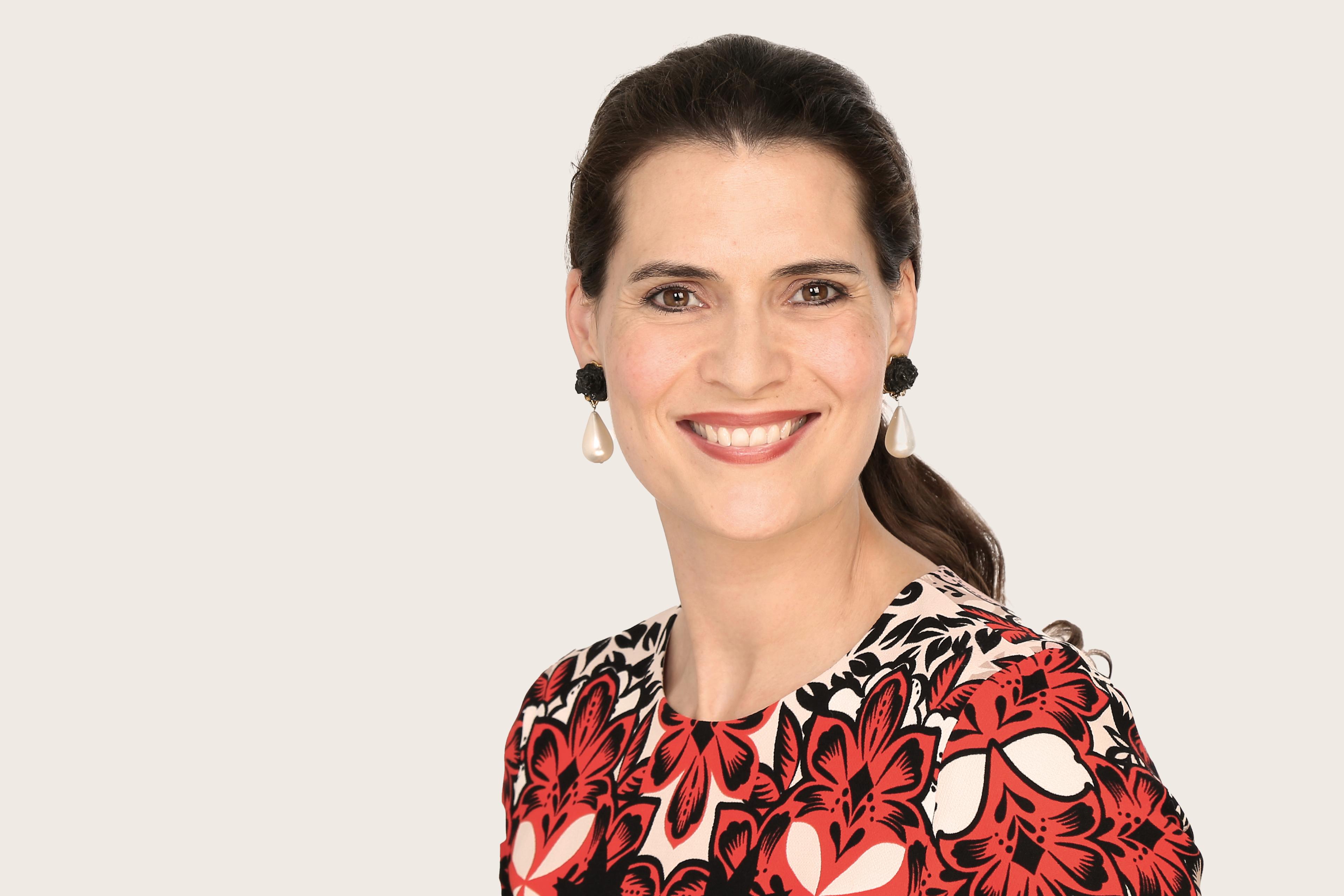 Janna Lena Baierle, eine Underwriter Art & Private Clients bei Hiscox, trägt  ein Kleid mit rot-schwarzem Blumenmuster und große Perlenohrringe; das lange Haar ist nach hinten gebunden.