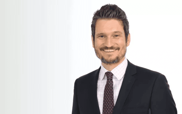timo kreitmair trägt einen dunklen Anzug, ein weißes Hemd und eine gemusterte Krawatte.
