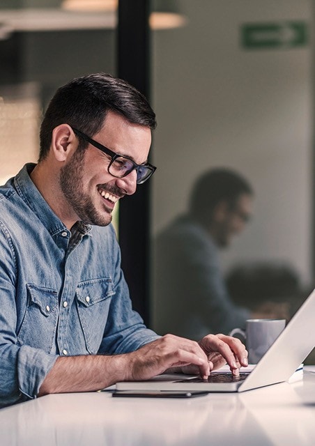 Ein junger Mann mit Brille und Jeanshemd arbeitet lächelnd am Laptop