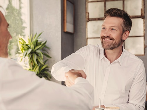 Zwei Personen in weißen Hemden geben sich in einem Innenraum die Hand; im Hintergrund sind ein Fenster mit mehreren Scheiben und eine Pflanze zu sehen.