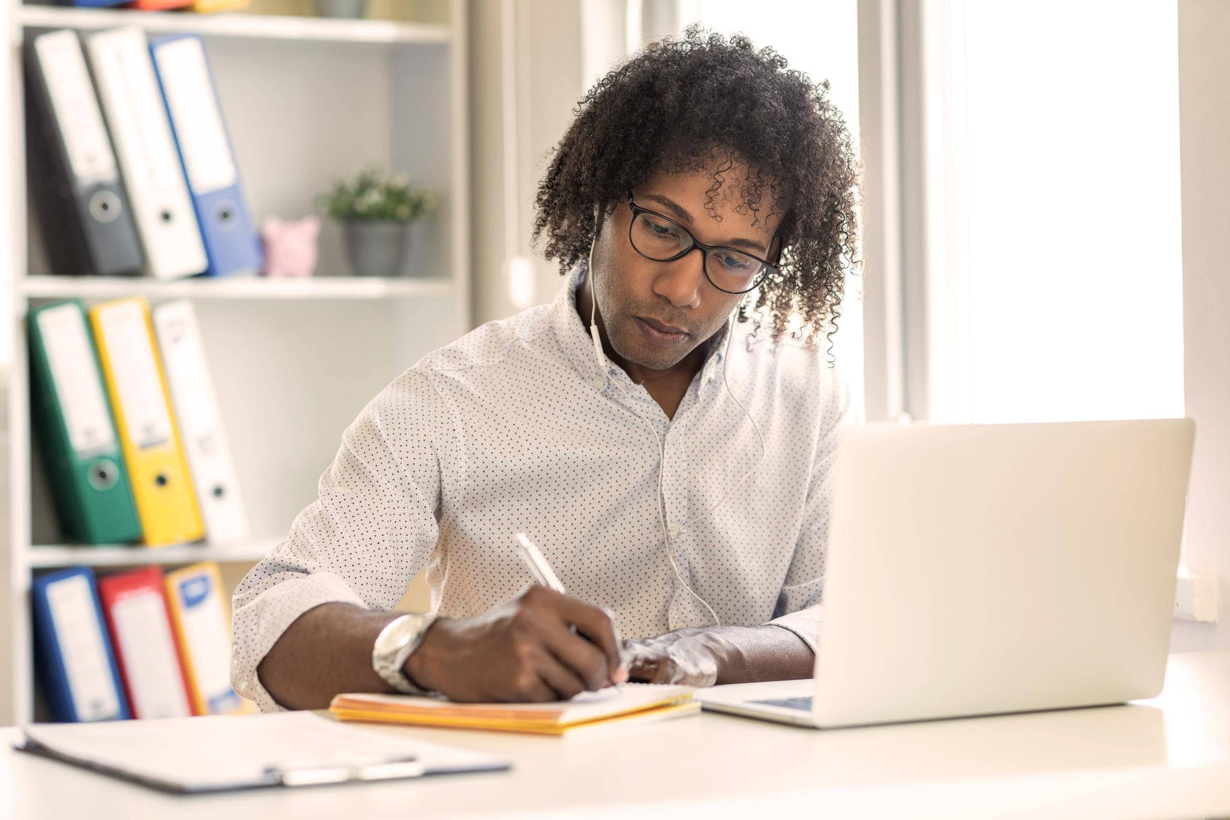 Ein Blogger mit Brille schreibt auf einem Blatt Papier, während er Kopfhörer trägt und an seinem Laptop arbeitet.
