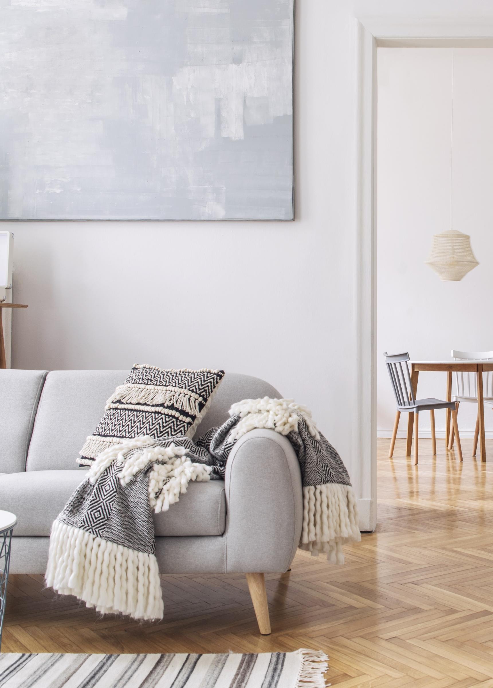 Ein modernes Wohnzimmer mit einem grauen Sofa, das mit zwei gemusterten Kissen und einer grauen Decke dekoriert ist. Im Hintergrund ist ein Esszimmer mit einem Holztisch und vier Stühlen zu sehen. Die Wände sind weiß gestrichen, und der Boden besteht aus hellem Holzparkett. An der Wand über dem Sofa hängt ein großes abstraktes Gemälde in Grautönen