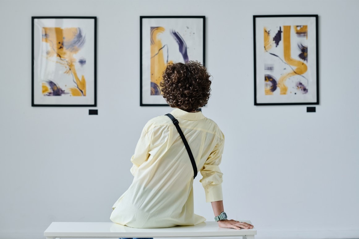 Eine Person sitzt auf einer Bank in einer Kunstgalerie und betrachtet drei abstrakte Gemälde an der Wand. Die Gemälde zeigen eine Mischung aus Gelb, Schwarz und Weiß mit verschiedenen Pinselstrichen und Formen. Die Person trägt ein helles Hemd und hat lockiges Haar