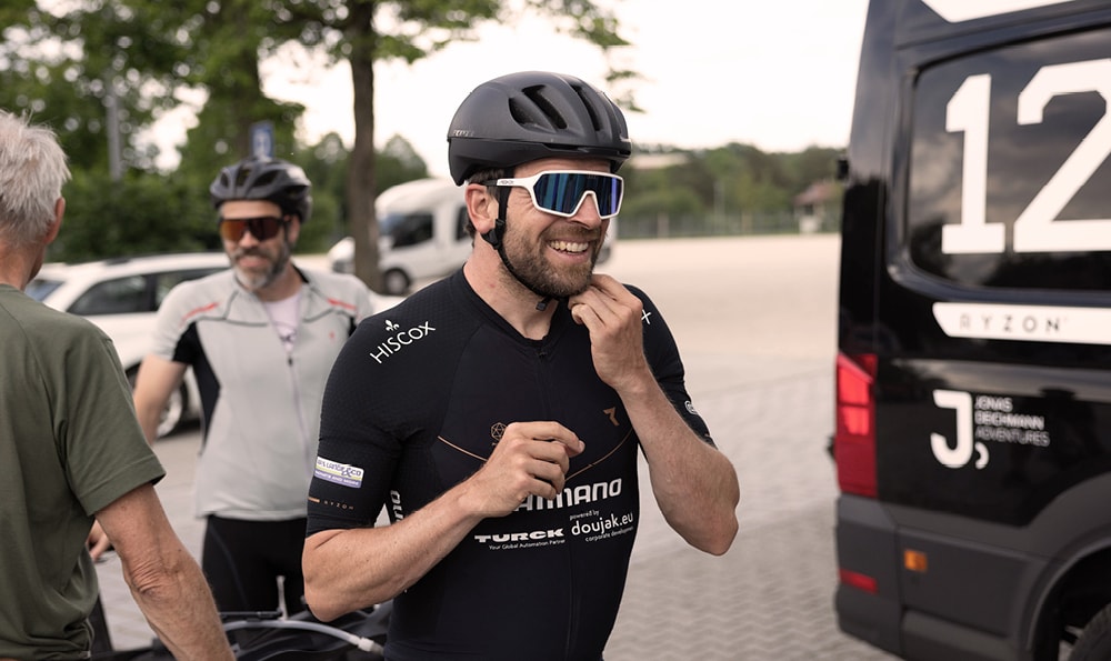 Jonas Deichmann, Leistungssportler aus Deutschland, steht in Fahrradmontur mit Hiscox Logo und Helm auf einer Straße. Zwei weitere Männer und ein Van befinden sich im Bild