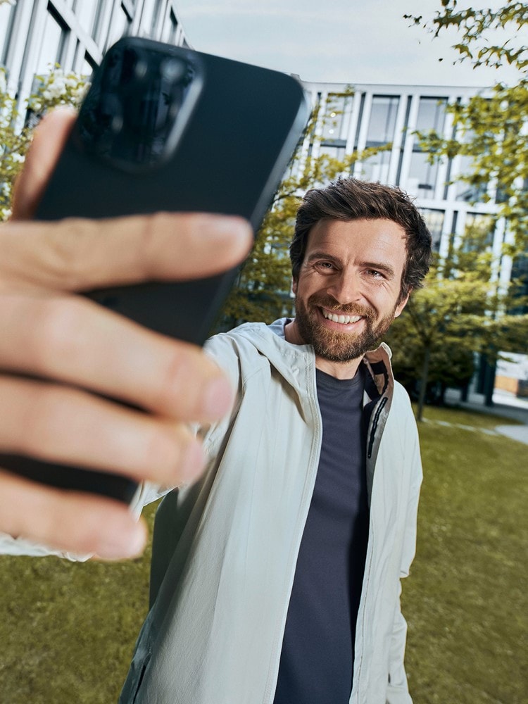 Das Bild zeigt eine Person, die ein Smartphone in einer ausgestreckten Hand hält, wahrscheinlich um ein Selfie zu machen. Im Hintergrund ist ein modernes Gebäude mit großen Fenstern und etwas Grün zu sehen, was auf eine Außenaufnahme hinweist.