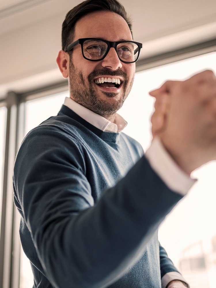 Junger Geschäftsmann mit dunklen Haaren und Brille in Anzug freut sich und schüttelt die Hand einer anderen Person.