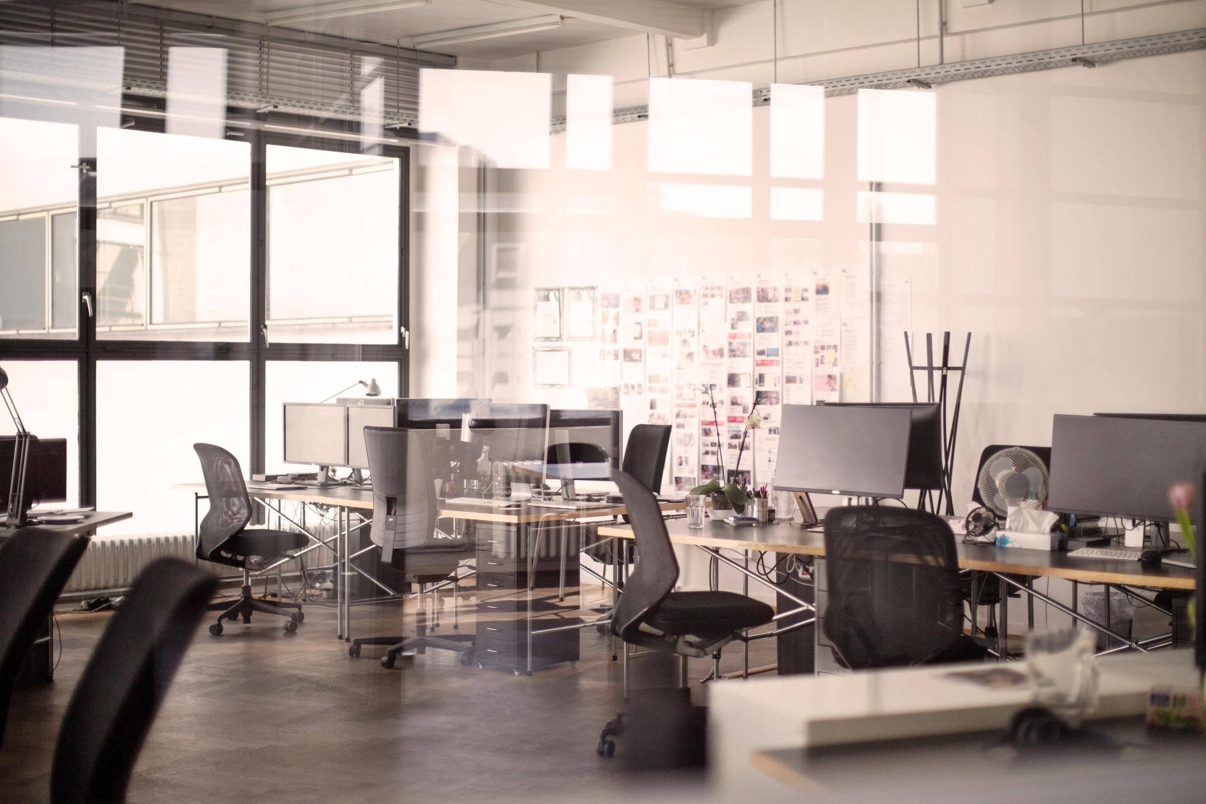 Ein modernes, helles Büro mit mehreren Schreibtischen und Computern. Große Fenster lassen viel Tageslicht herein. An den Wänden hängen Bilder und Notizen.