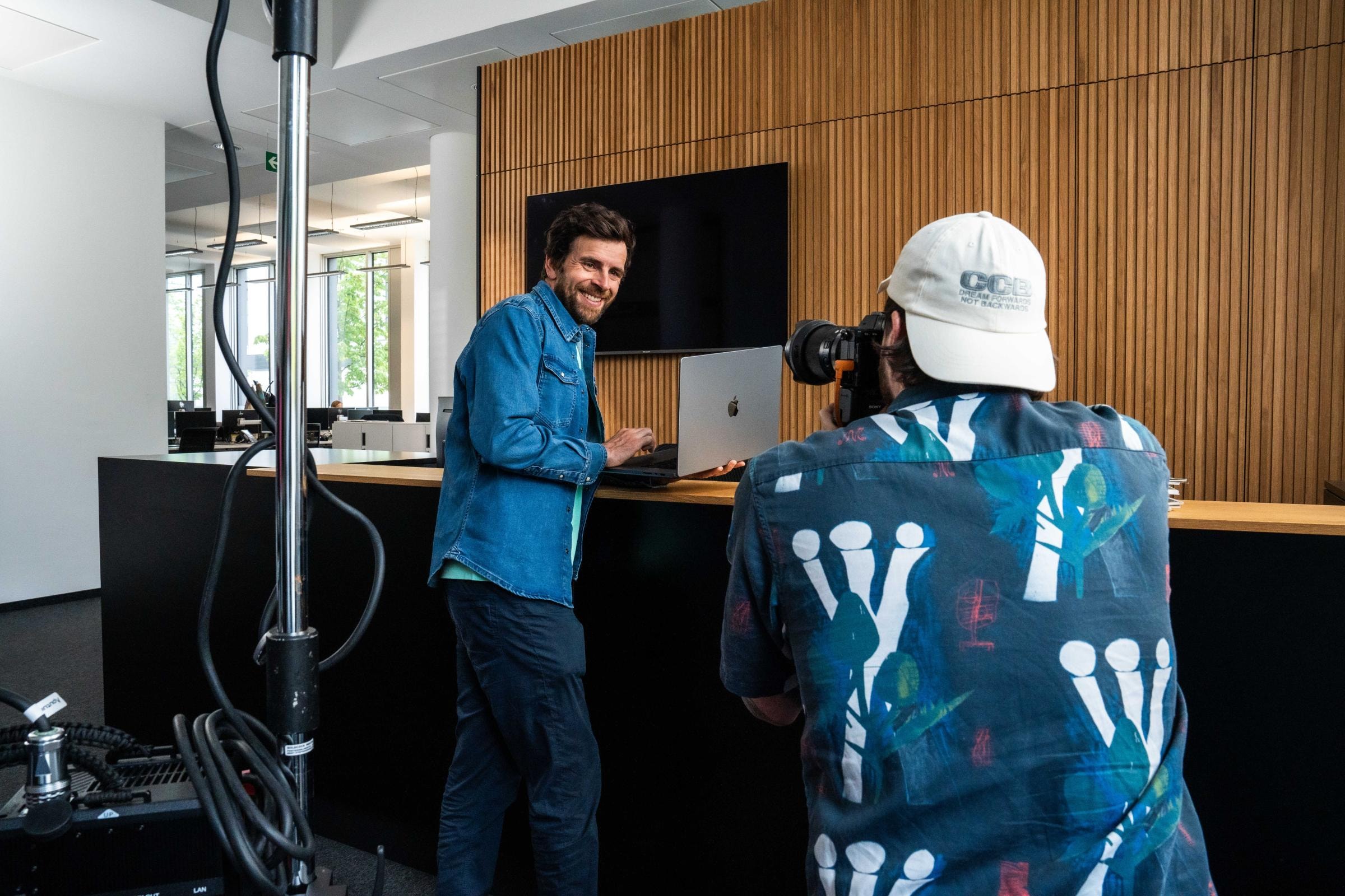 Ein Fotograf mit einer weißen Kappe und einem bunten Hemd fotografiert eine Person, die vor einem Schreibtisch steht. Die Person trägt eine blaue Jeansjacke und schaut auf einen Computerbildschirm.