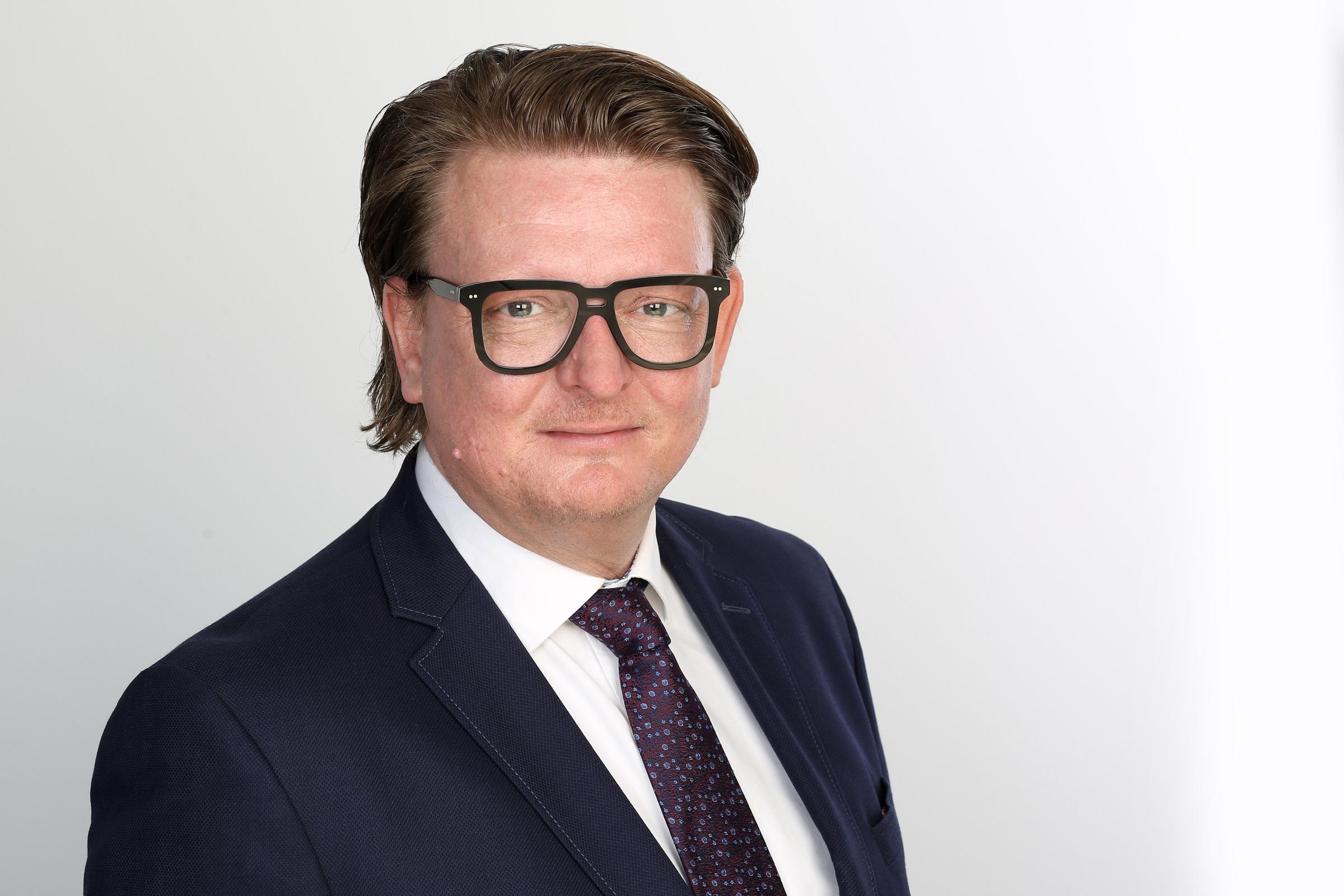 Marc Thamm trägt einen dunklen Anzug, ein weißes Hemd und eine dunkle Krawatte mit kleinen Mustern.