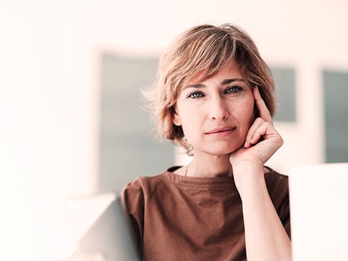 Eine Person mit kurzen Haaren trägt ein braunes Hemd, und die Person hält eine Hand an die Seite ihres Gesichts.