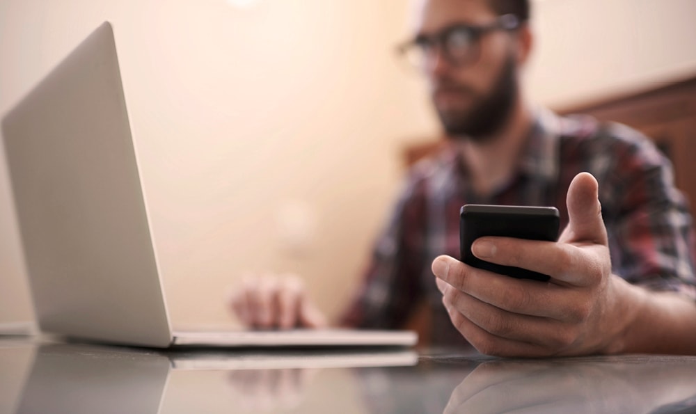 Eine Person sitzt an einem Schreibtisch, benutzt einen Laptop und hält ein Smartphone. Das Gesicht der Person ist unscharf, und der Fokus liegt auf den verwendeten Geräten.