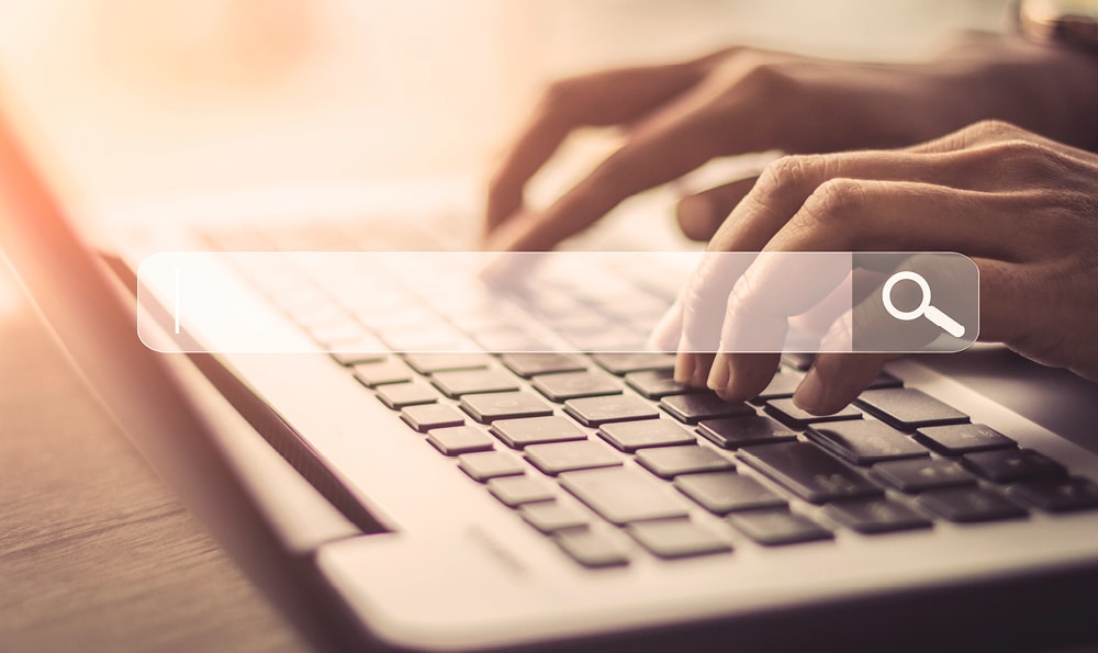  Eine Nahaufnahme von Händen, die auf einer Laptop-Tastatur tippen und in der Mitte des Bildes ist eine Suchleiste mit einem Lupensymbol zu sehen