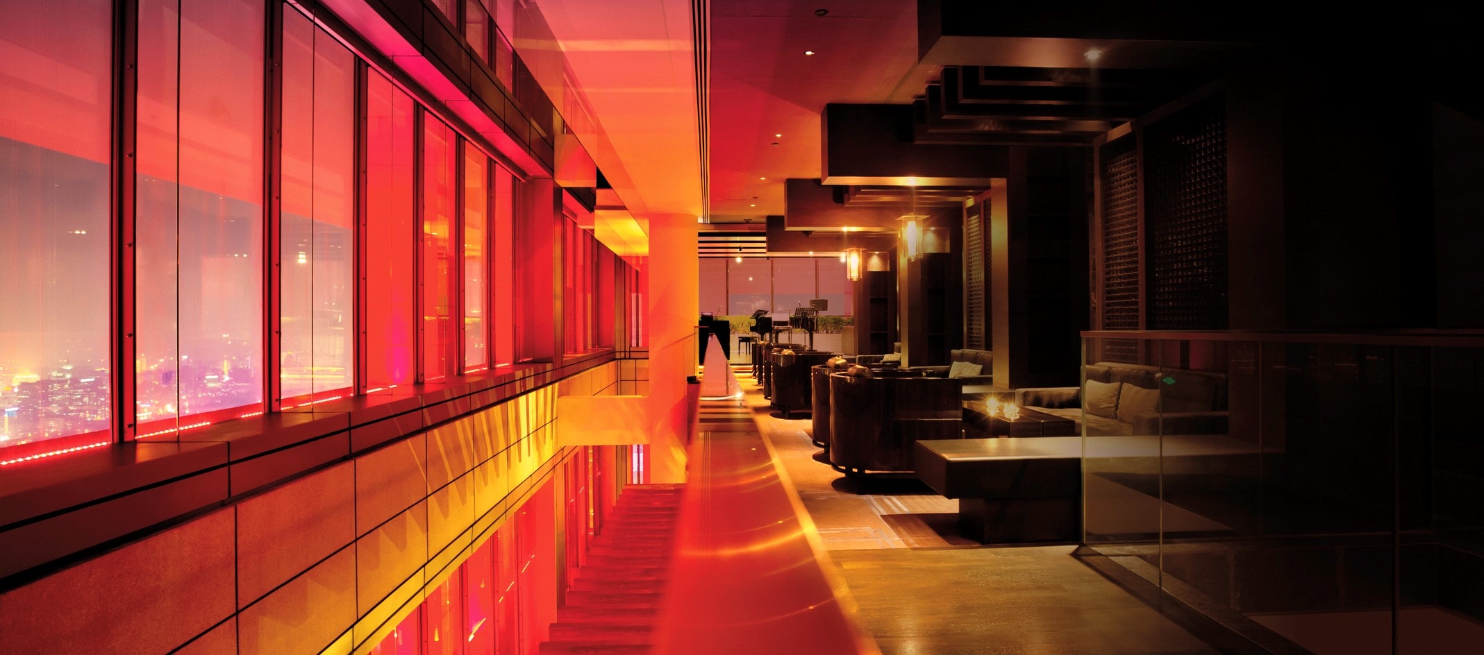 Ein modernes, stilvolles Interieur mit einer langen, beleuchteten Passage. Links sind große Fenster mit rotem Licht, das die Stadtlandschaft draußen zeigt. Rechts befinden sich Sitzbereiche mit Sofas  und Tischen unter warmem Licht. Eine Person steht am Ende der Passage und schaut nach draußen
