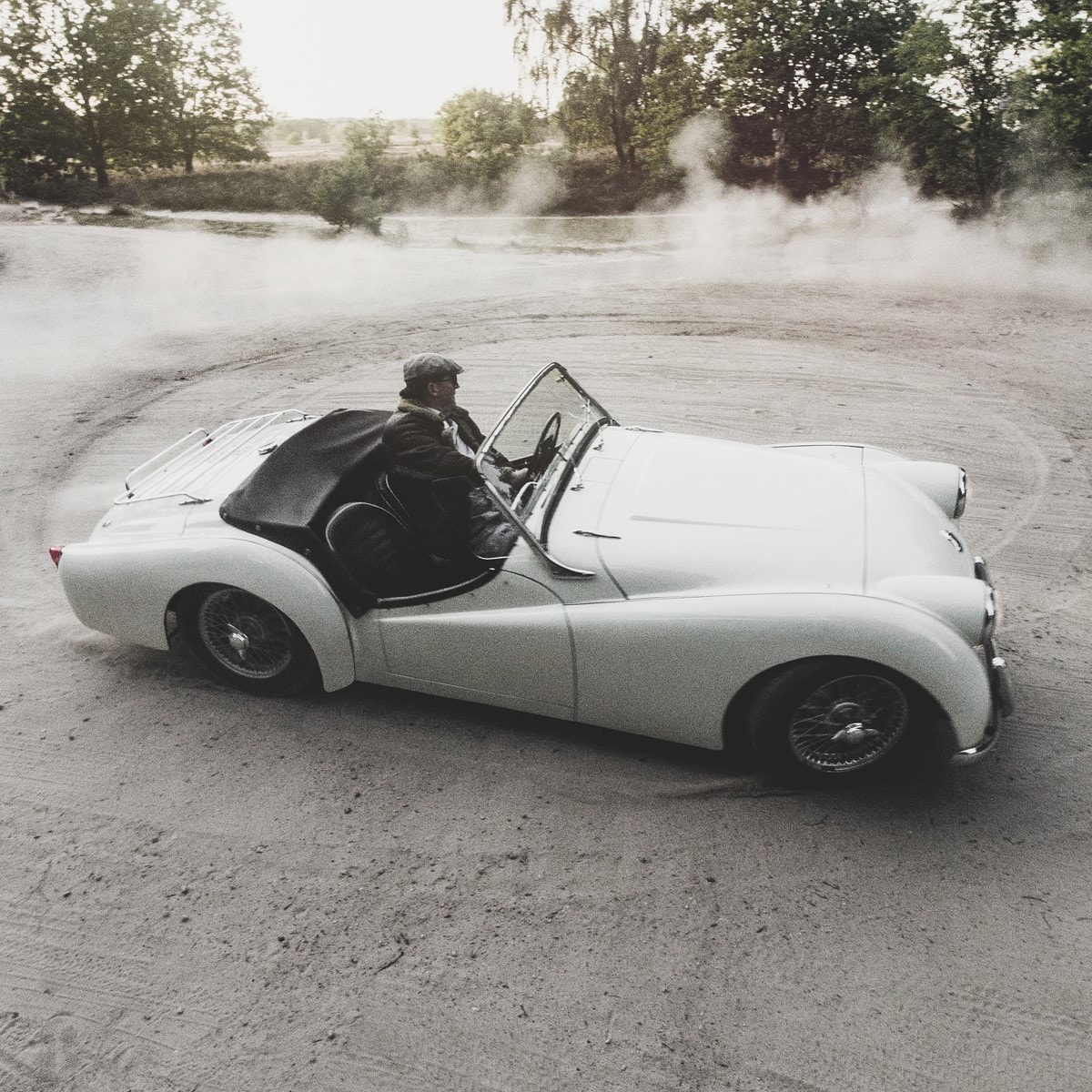 Weißes Cabrio-Oldtimer fährt auf staubiger Straße, der Fahrer trägt Mütze und Jacke. Im Hintergrund Bäume und aufgewirbelter Staub