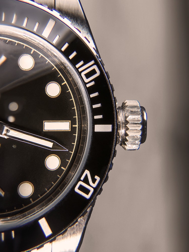 Eine Nahaufnahme einer Armbanduhr mit schwarzem Rand und Zifferblatt, weißen Markierungen und leuchtenden Punkten. Die Krone ist auf der rechten Seite sichtbar.