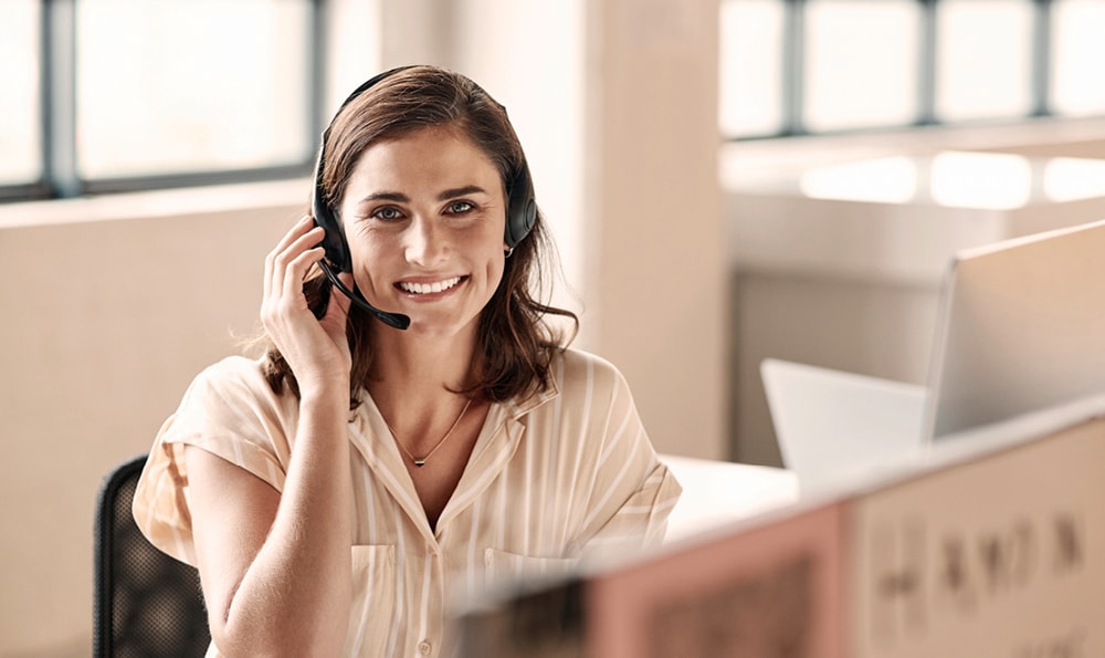 Junge Frau lächelt in die Kamera. Sie trägt ein Headset mit Micro und eine helle Bluse.