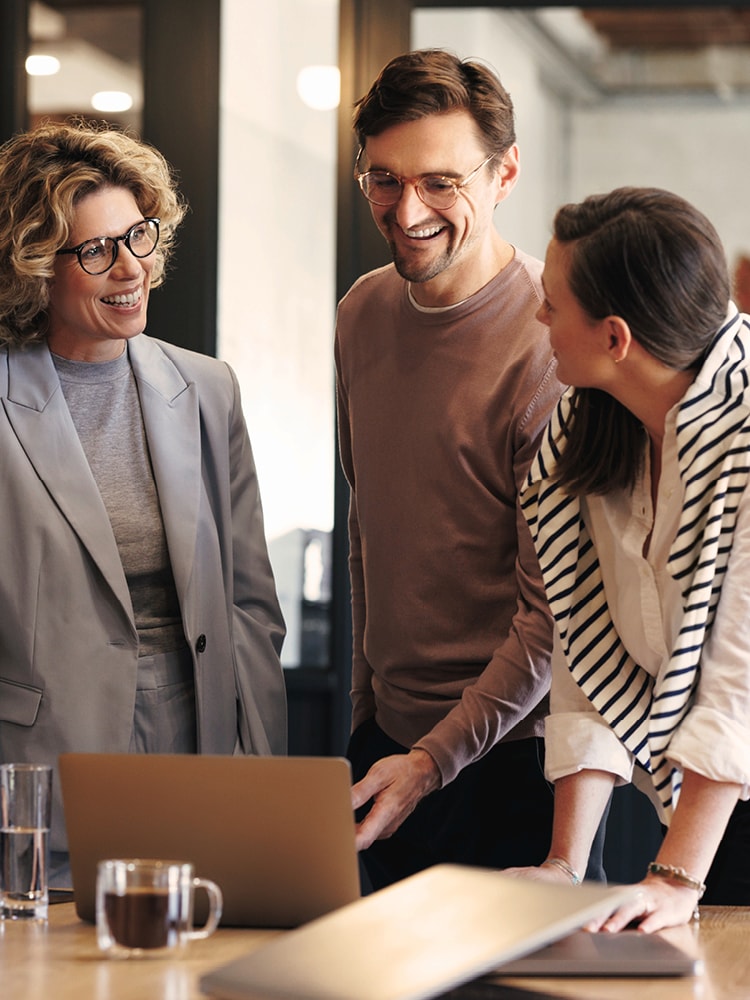 Drei Personen stehen um einen Tisch mit einem Laptop. Eine Person zeigt auf den Bildschirm. Auf dem Tisch stehen außerdem ein Glas Wasser und eine Tasse Kaffee. Im Hintergrund ist ein Büro mit Glaswänden und Beleuchtung zu sehen.