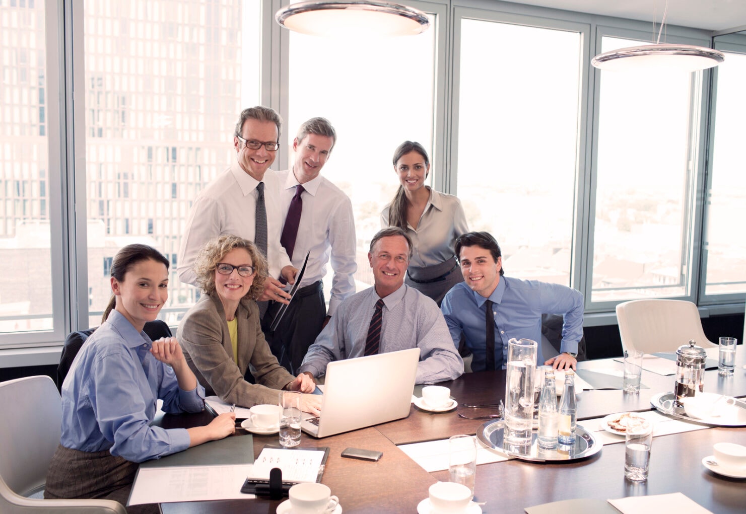 Sieben lächelnde Geschäftsleute sitzen um einen Laptop an einem Meetingtisch in Business-Kleidung.