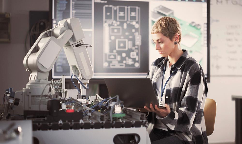 Eine junge blonde Frau mit kurzen Haaren sitzt vor einer Werkbank mit elektronischen Geräten und einem Roboterarm. Sie hält einen Laptop in der Hand über ihrem Schoß, in den sie hineinschaut. Im Hintergrund sieht man verschwommen einen großen Bildschirm mit technischen Zeichnungen.