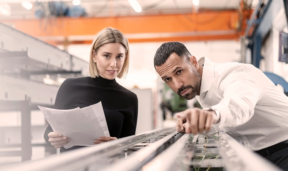 Eine blonde Frau mit kinnlangem Haar und schwarzem Rollkragenpullover und ein Mann mit dunklem Haar, Bart und in einem weißen Hemd stehen in einer Fertigungshalle an einer langen flachen technischen Konstruktion mit ein paar Kabeln. Die Frau hält drei weiße Blätter mit Aufzeichnungen in den Händen. Der Mann zeigt entlang der Konstruktion in unsere Richtung, in die beide interessiert schauen.