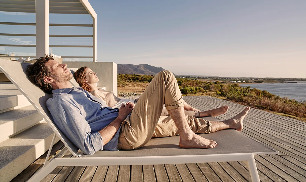 Ein Mann und eine Frau liegen mit geschlossenen Augen auf einer Sonnenliege auf der Terrasse eines Ferienhaues. Im Hintergrund sieht man Dünnen und das Meer