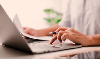 Ein Unbekannter arbeitet am Laptop und hält in einer Hand ein Papier und in der anderen einen Stift