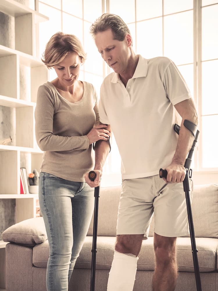 Eine blonde Frau stützt einen blonden Mann, der an Krücken geht und einen verbundenen Unterschenkel hat