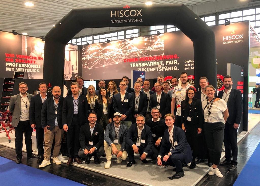 Eine Gruppe von Menschen posiert für ein Foto bei einer Veranstaltung oder Ausstellung. Sie stehen unter einem großen schwarzen Torbogen mit der Aufschrift 'HISCOX WISSEN VERSICHERT'.