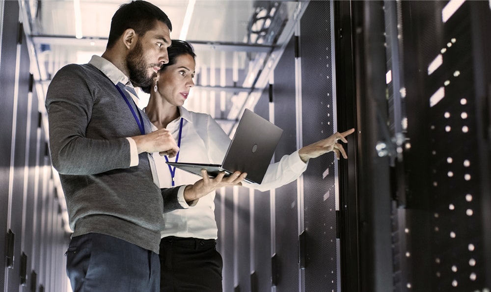 Zwei Personen in einem Serverraum, eine Person hält einen Laptop und die andere zeigt auf ein Server-Rack. Beide tragen ID-Karten um den Hals.