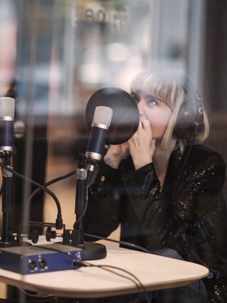 Frau mit blonden Haare und Lederjacke sitzt vor eine Podcast-Mikrofone und trägt Kopfhörer.