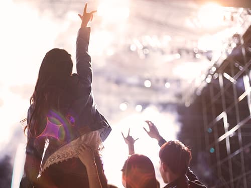 Menschenmenge bei einer Live-Veranstaltung: Eine Person sitzt auf den Schultern einer anderen und hebt jubelnd die Arme vor einer hell erleuchteten Bühne – Stimmung von Konzert oder Event.