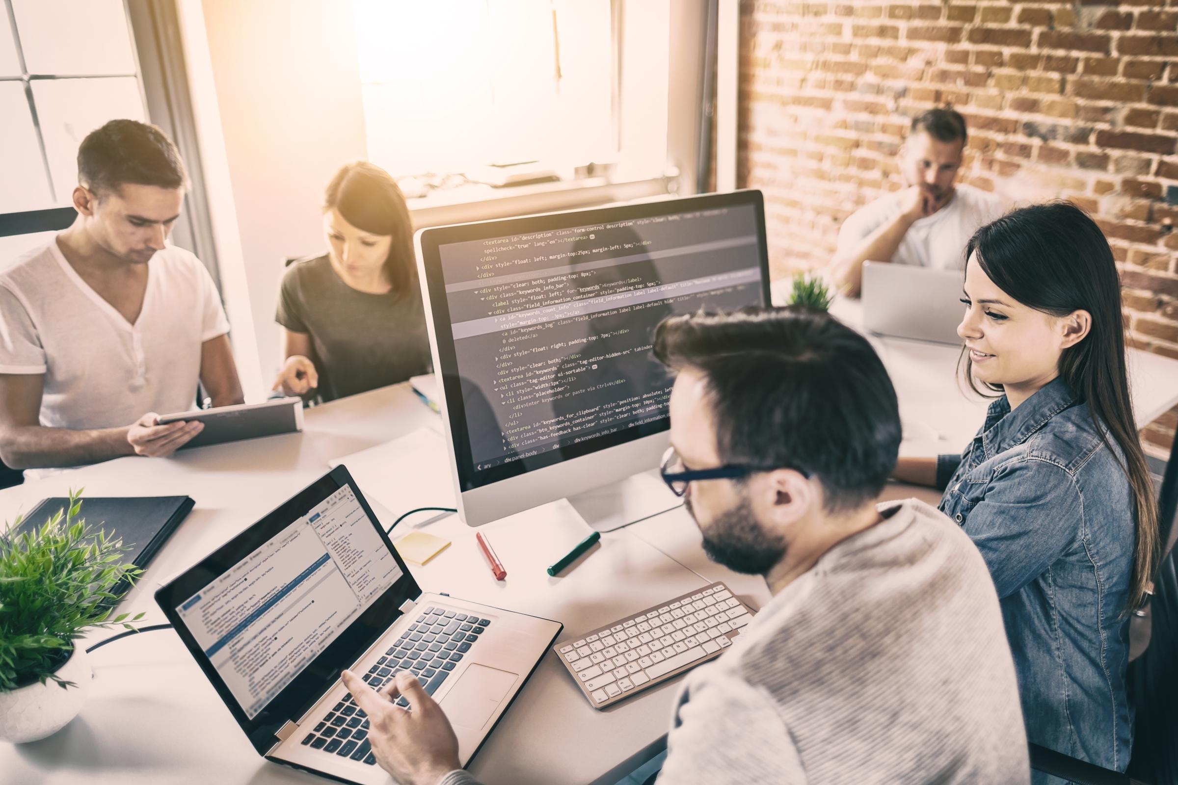 Gruppe von fünf Personen in einem Büro, arbeitend an Laptops und Monitoren mit Code; Gesichter unscharf, Ziegelwände und große Fenster im Hintergrund.