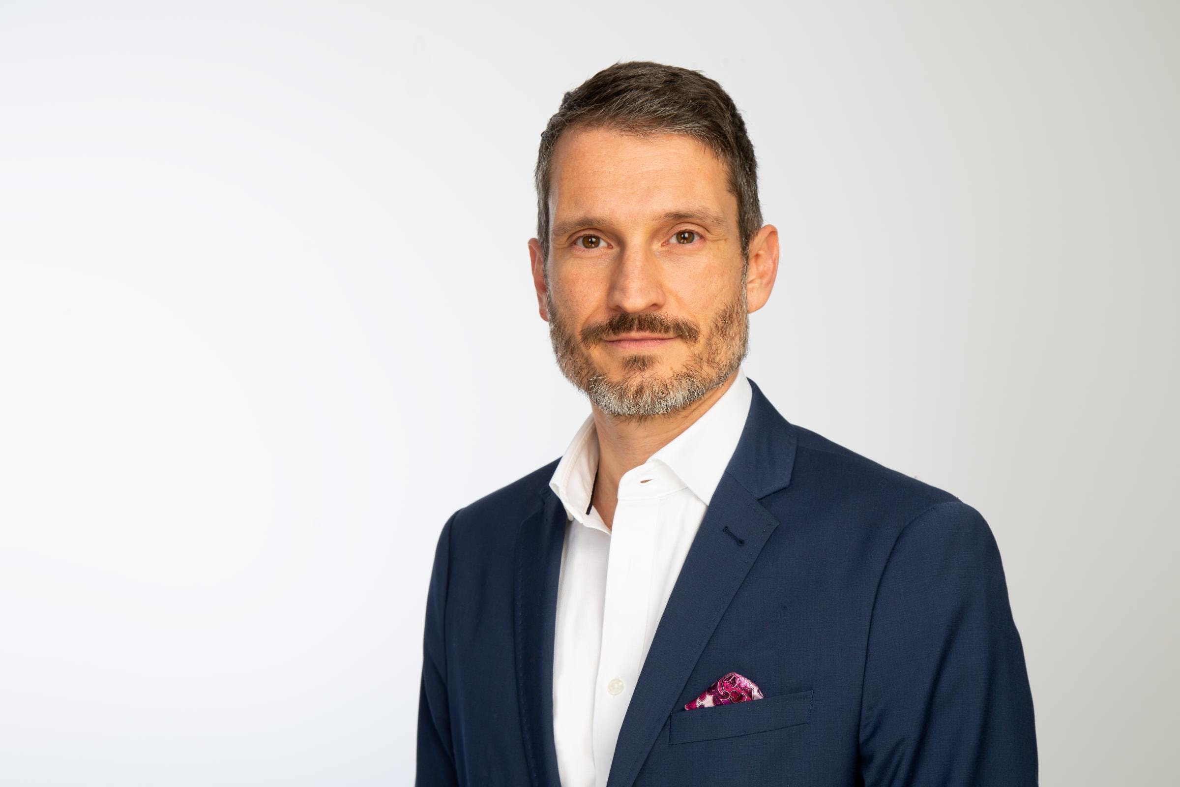 Timo Kreitmair trägt ein dunkles Sakko, ein weißes Hemd und blickt in die Kamera.