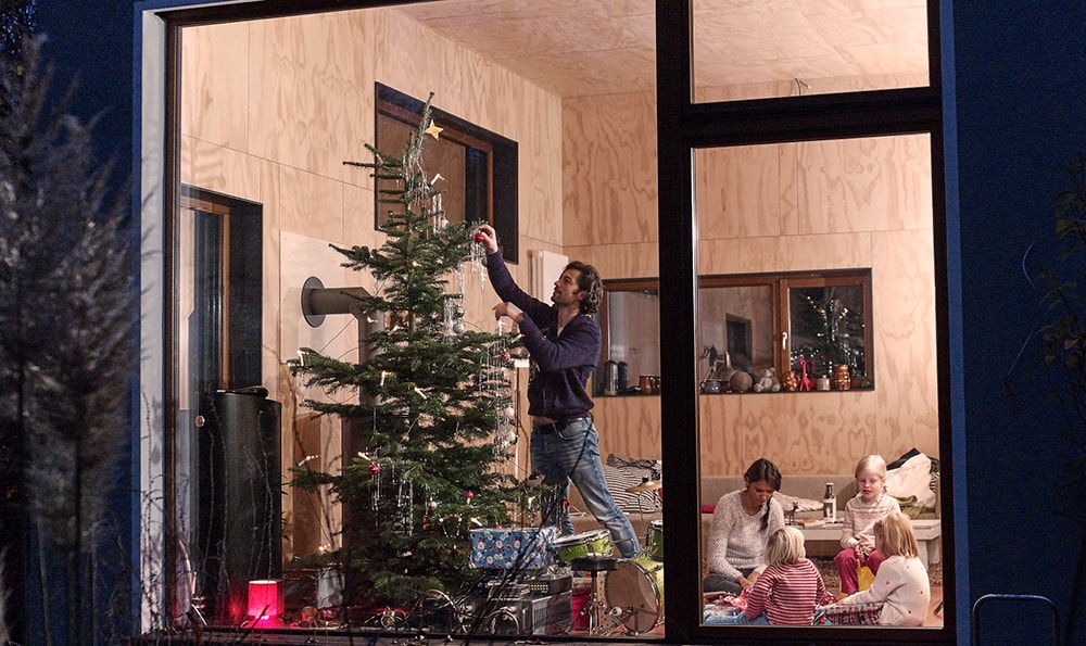 Eine Familie schmückt im Haus einen Weihnachtsbaum. Eine Person steht auf einem Stuhl und hängt eine Kugel auf, während drei weitere am Tisch sitzen. Draußen ist es dunkel.