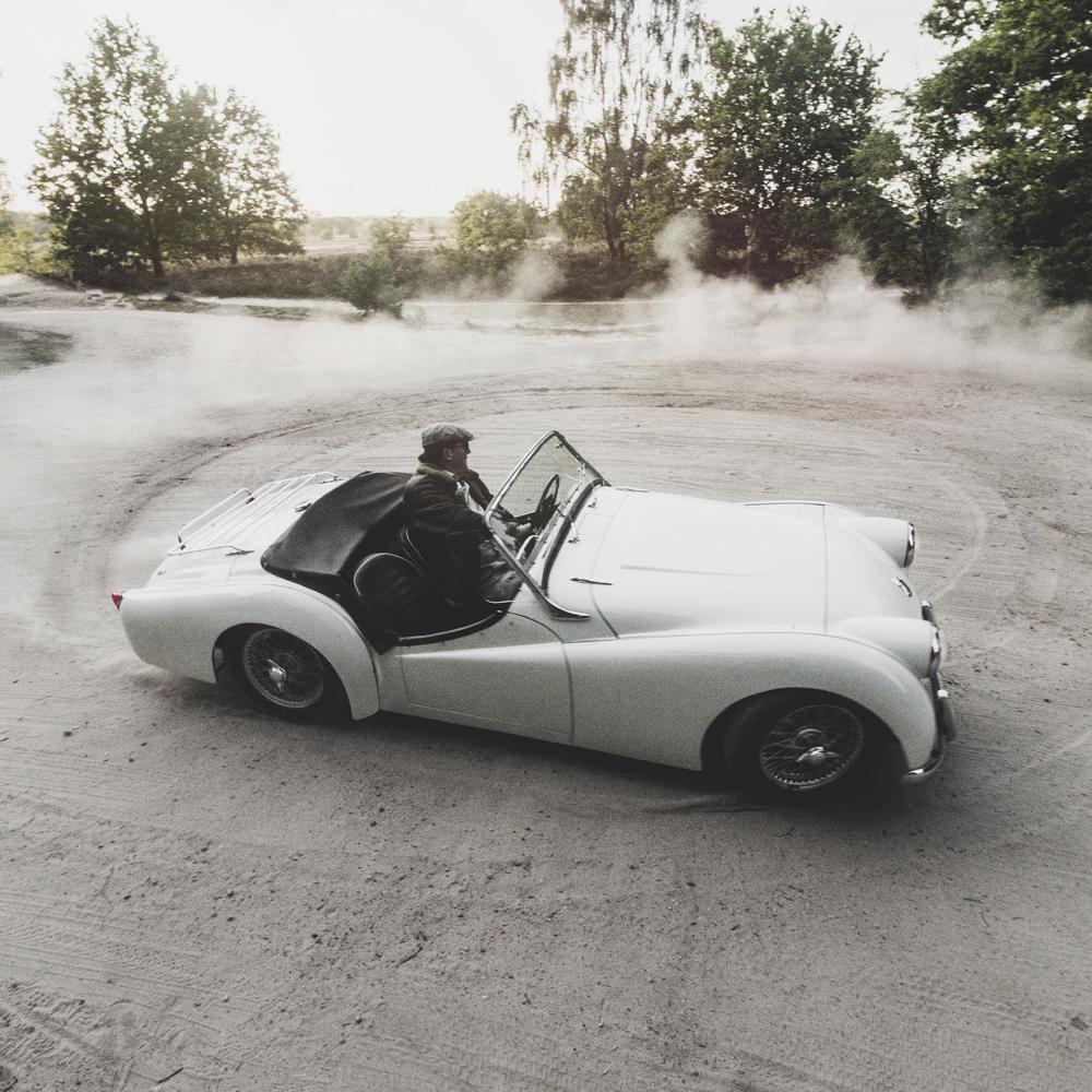 Ein weißes Cabrio-Oldtimer fährt auf einem Feldweg und wirbelt Staub auf. Der Fahrer trägt einen Hut. Im Hintergrund sind Bäume und Sonnenlicht zu sehen.