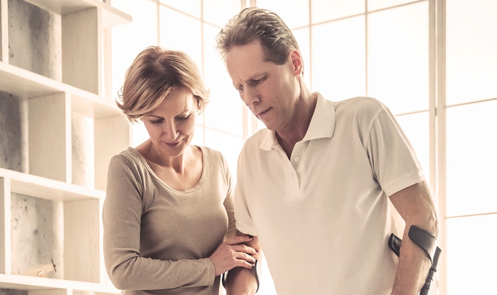 Ein Mann mit Krücken und eine Frau stehen nebeneinander in einem hellen Raum. Die Frau hält den Arm des Mannes unterstützend fest