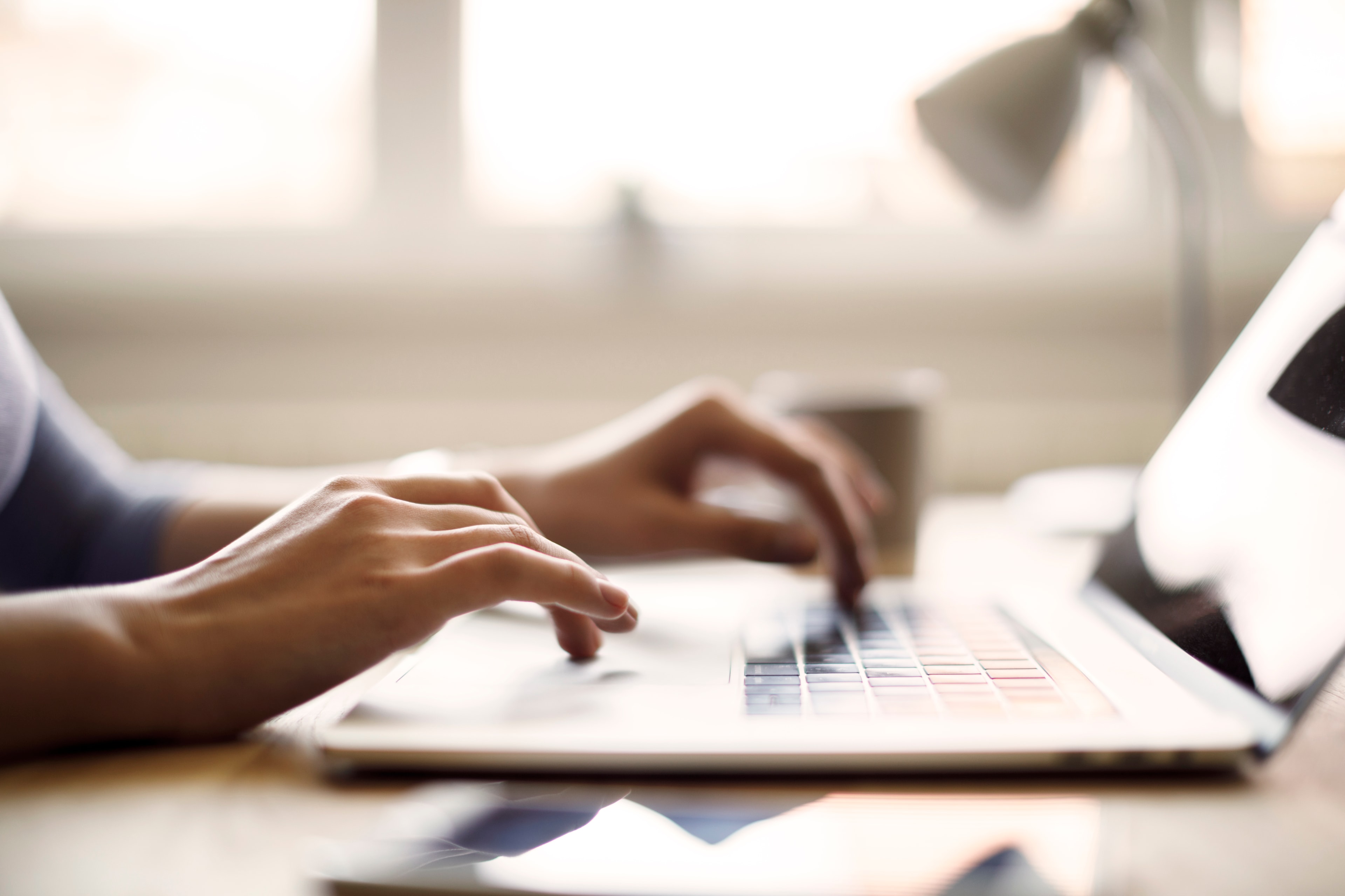 Eine Person tippt auf einer Laptop-Tastatur. Im Hintergrund sind ein Fenster, eine Lampe und eine Tasse zu sehen.