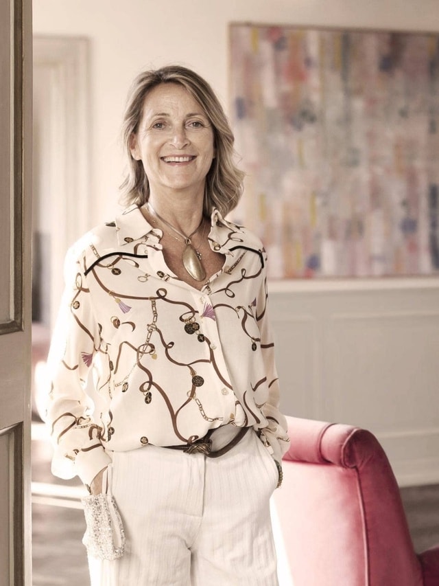 Eine Person steht in einem Raum mit hellen Wänden und Holzfußboden, trägt ein weißes Hemd mit Blattmuster und weiße Hose; im Hintergrund ein abstraktes Gemälde und ein roter Stuhl.