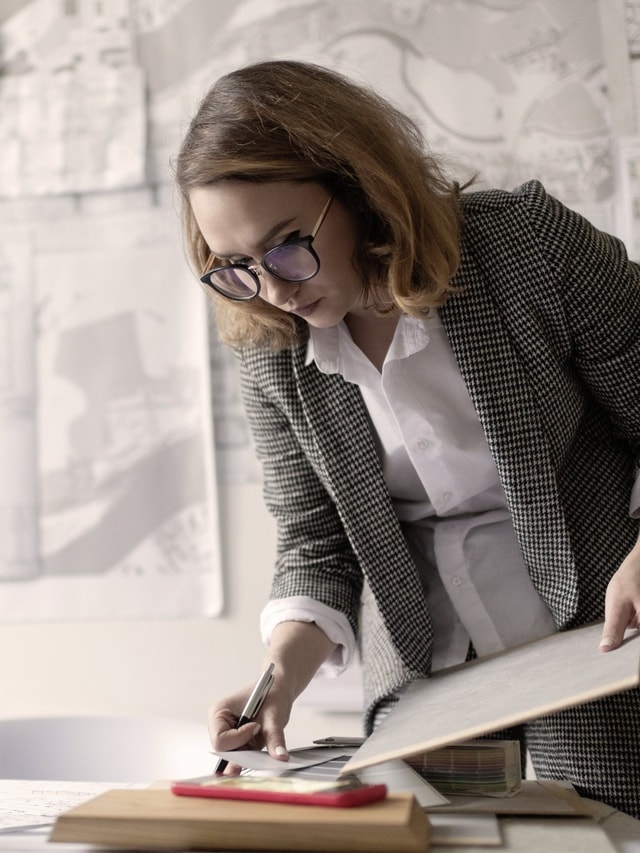 Eine Frau mit Brille und kariertem Blazer arbeitet konzentriert an einem Projekt am Schreibtisch.