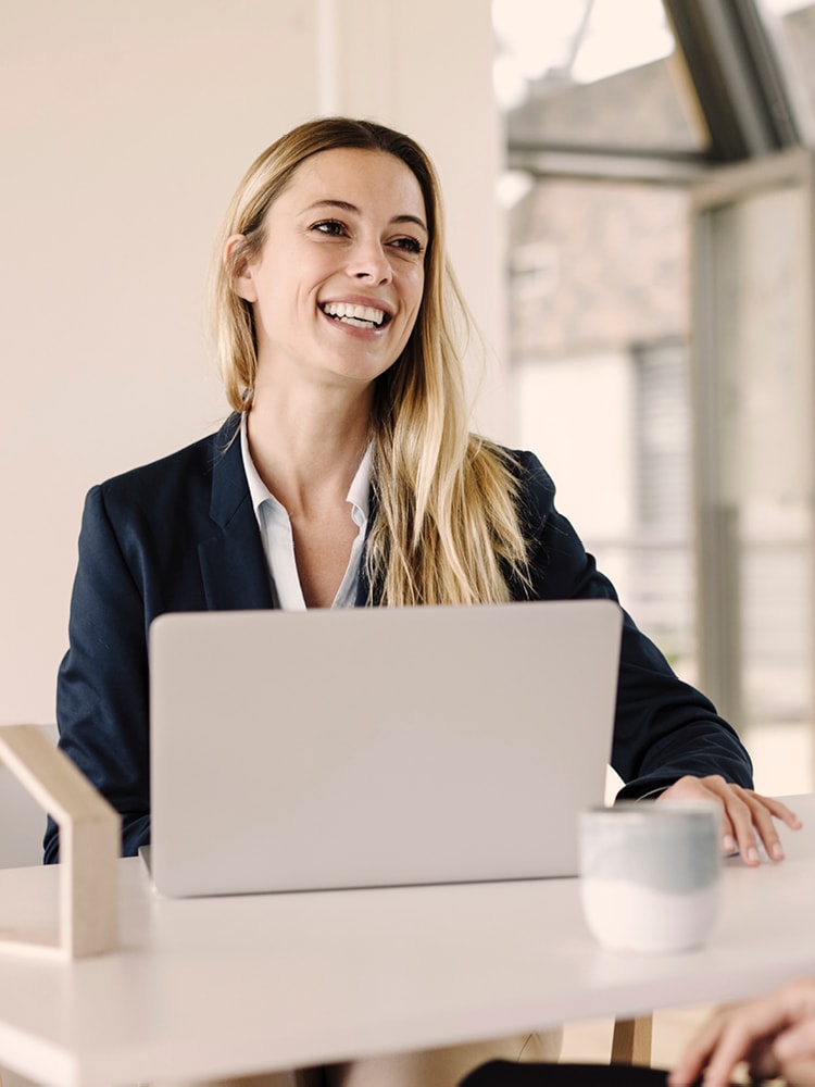 Person mit langen blonden Haaren und dunklem Blazer sitzt vor einem Laptop an einem Schreibtisch; Gesicht unscharf, im Hintergrund große Fenster mit Tageslicht.