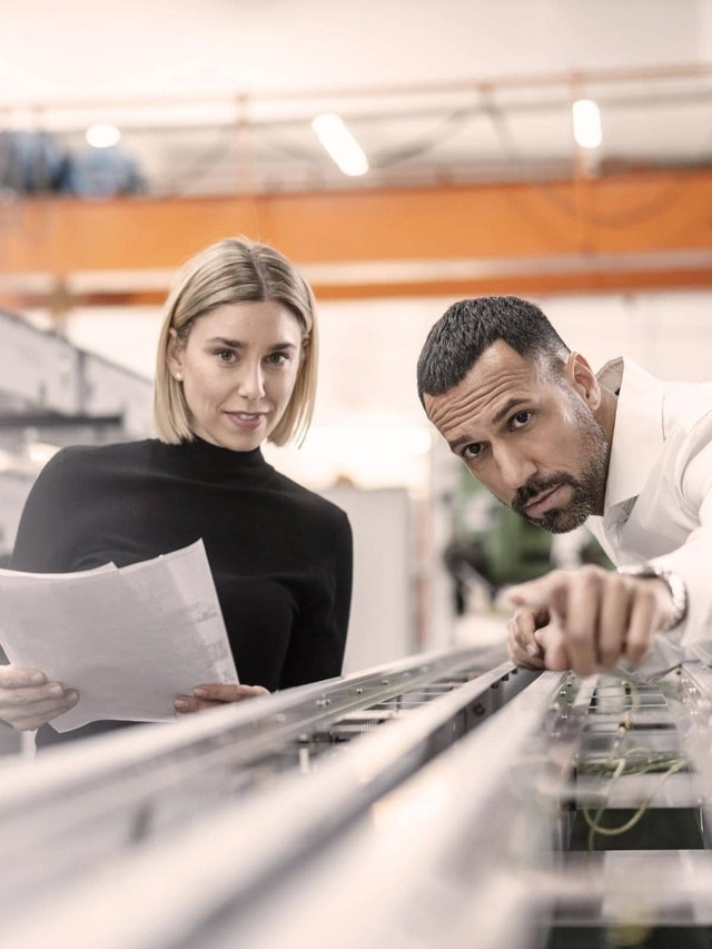 Zwei Personen in einer industriellen Umgebung. Eine Person hält Papiere, die andere zeigt auf eine Maschine. Im Hintergrund sind Fabrikelemente zu sehen.