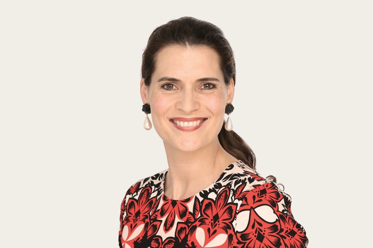 Janna Lena Baierle, eine Underwriter Art & Private Clients bei Hiscox, trägt ein Kleid mit rot-schwarzem Blumenmuster und große Perlenohrringe; das lange Haar ist nach hinten gebunden.