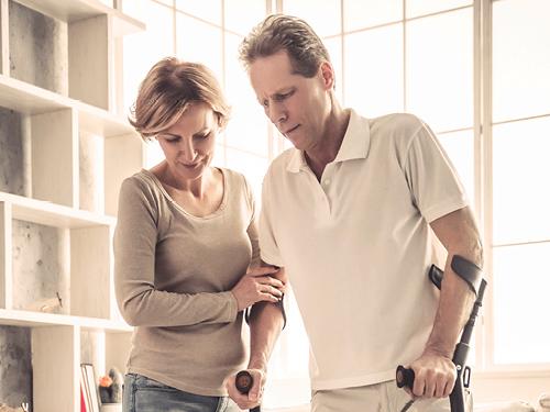 Zwei Personen stehen nah beieinander. Eine Person hält eine Krücke und scheint Unterstützung von der anderen Person zu erhalten, die einen Arm um den Rücken der ersten Person gelegt hat und deren Hand hält. Im Hintergrund ist ein großes Fenster mit natürlichem Licht und einige Regale auf der linken Seite des Bildes zu sehen.