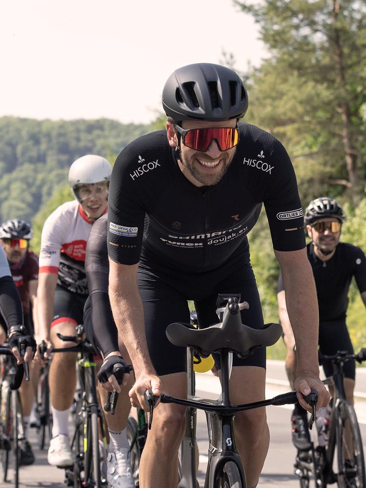 Eine Gruppe von Radfahrern fährt auf einer Straße; im Vordergrund ein Fahrer in schwarzer Kleidung mit „HISCOX“-Logo auf den Ärmeln, im Hintergrund weitere Radfahrer und grüne Bäume entlang der Strecke.